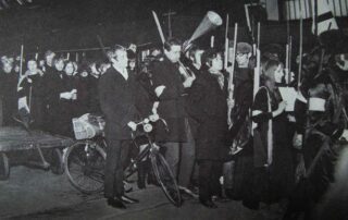 The Russian Revolution by Bradford Art College Theatre Group, 1967. A large group of men and women in long black coats hold weapons, loud speakers, one has a bike, some are carrying papers. A march.