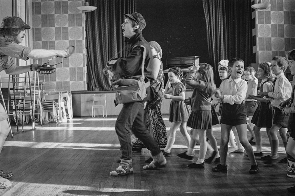 Theatre in Education from The Belgrade Theatre, 1960s (Photo: Tony Baker). A school hall. An adult male leads a pack of school children with happy faces towards another man holding a toy tank. One prominent child is howling with laughter.