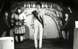 Bearing the Weight by Spare Tyre, 1979. Black and white photo of a performance inwhich a woman in a white is stood in front of a psychadelic backdrop with three two-dimensional women, one is in big furry clothes and the other two are mirror images of one another, they are a dancer in blacks.