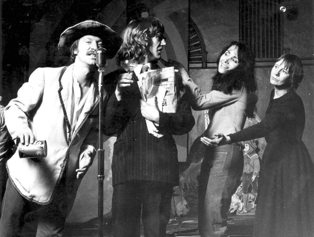 7:84’s The Cheviot, The Stag and the Black, Black Oil, 1973. 4 performers on stage, two men two women. On stage right the two men, one speaks into a vintage microphone in a cheaper suit and the one next to him is in a more expensive suit holding a wad of newspapers. The two women are both communicating with the man in the more expensive suit, one with a hand on the shoulder warmly, the other with a hand outstretched expectantly.