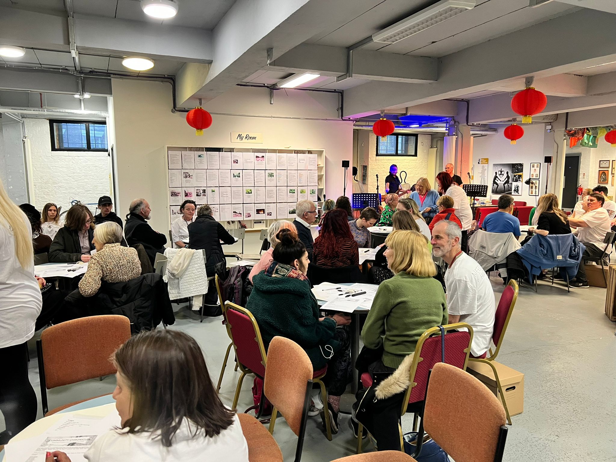 A bright white and grey room full of people who are sat around round tables waiting for a performance to begin