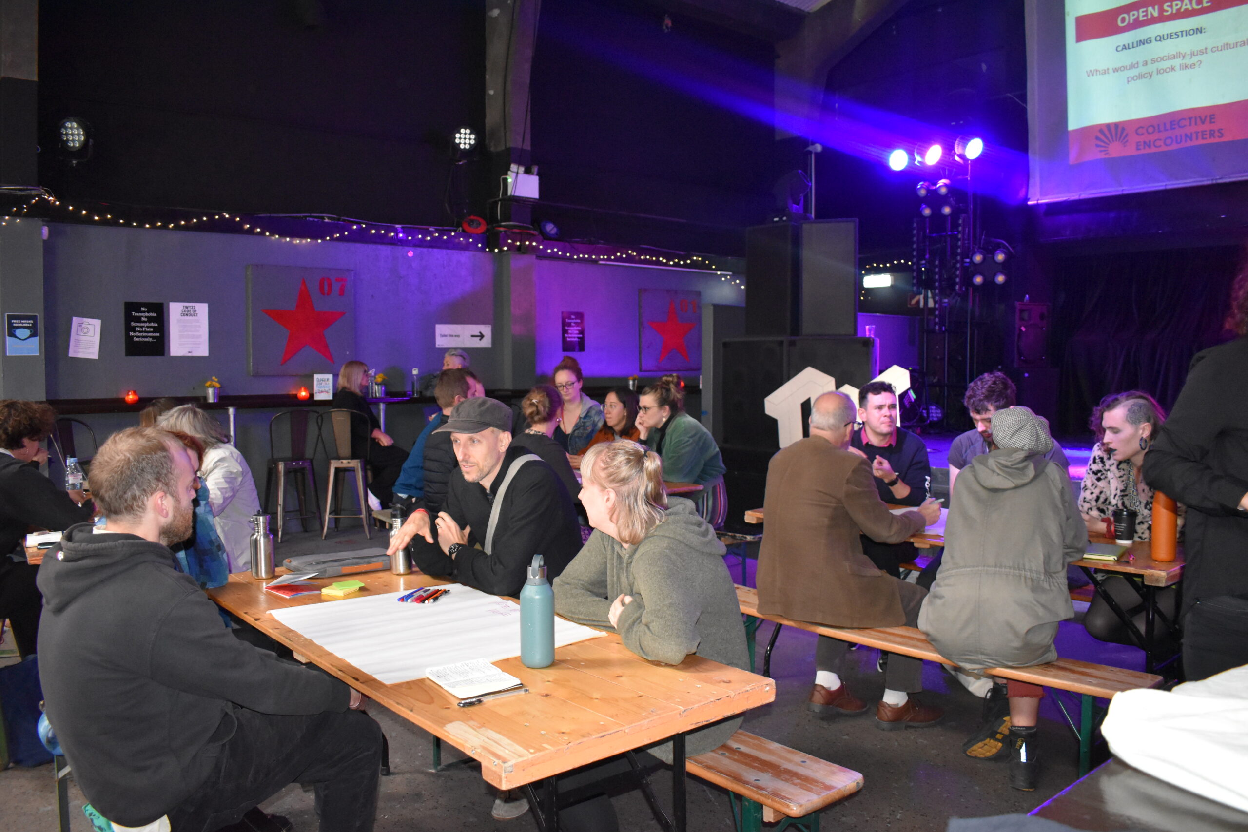 A dark room with purple lights. People are sitting around long wooden bench tables writing on large paper