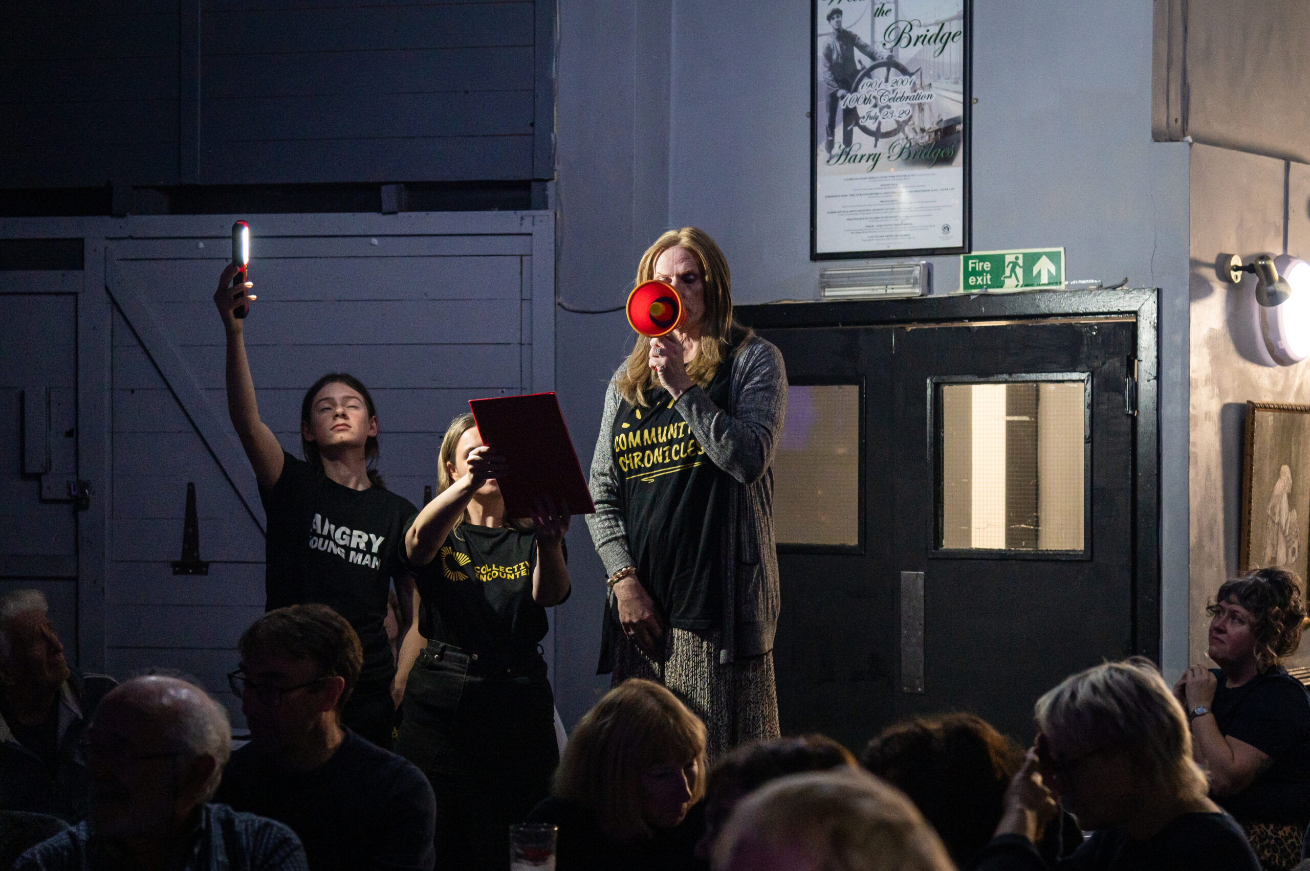 A performer is standing on a soapbox speaking into a megaphone, while another performer holds a board up for them to read and another holds a light on them